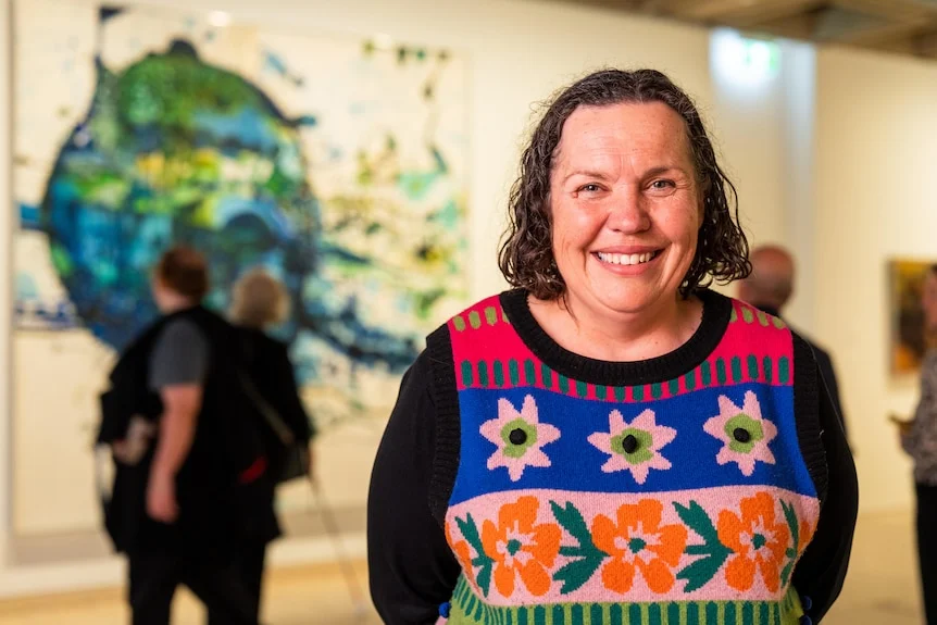 Deborah Bennetts in a black long sleeve, colourful knit vest, brown hair, smiling inside a gallery.