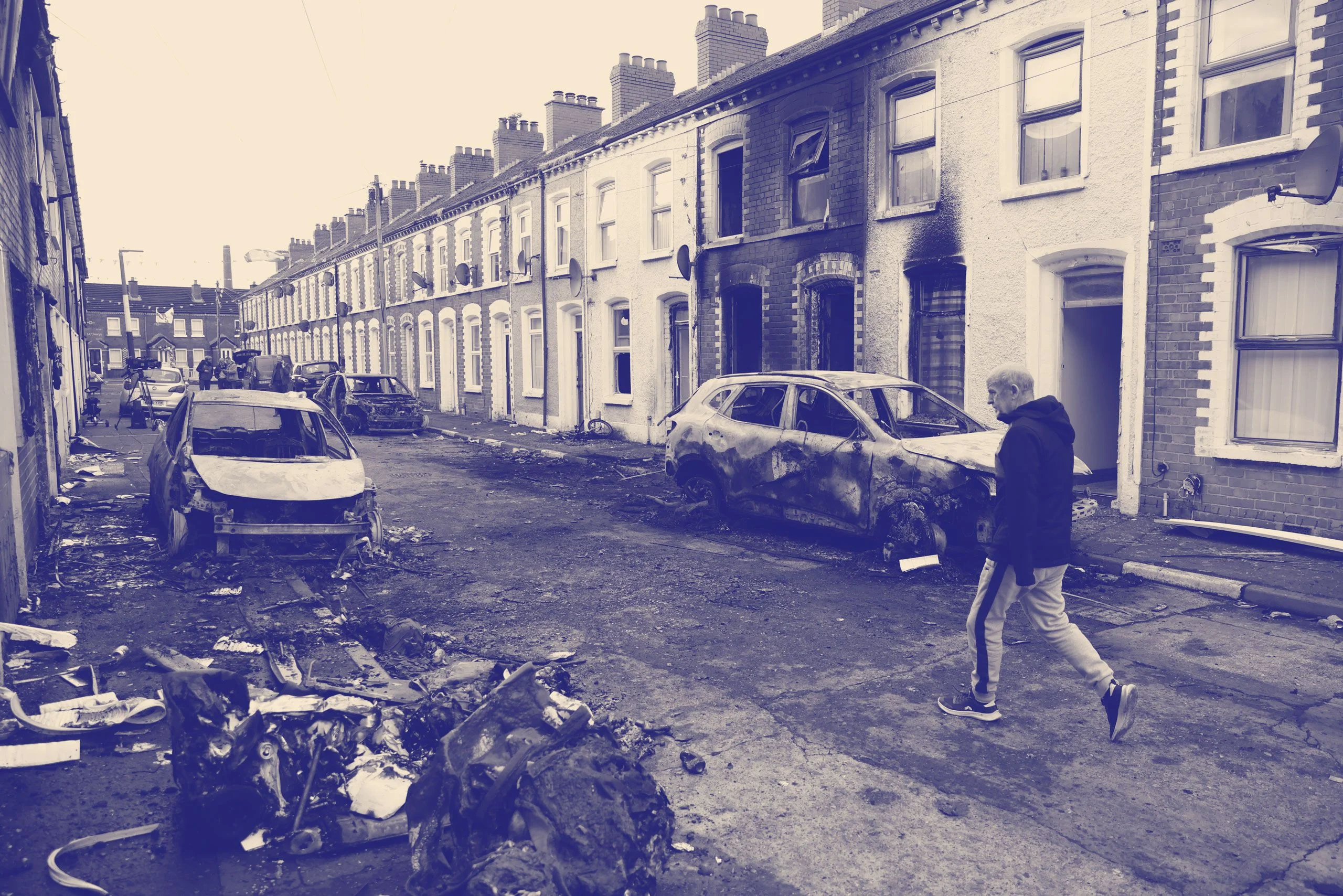 A man walks down a street lined with burned buildings and cars.
