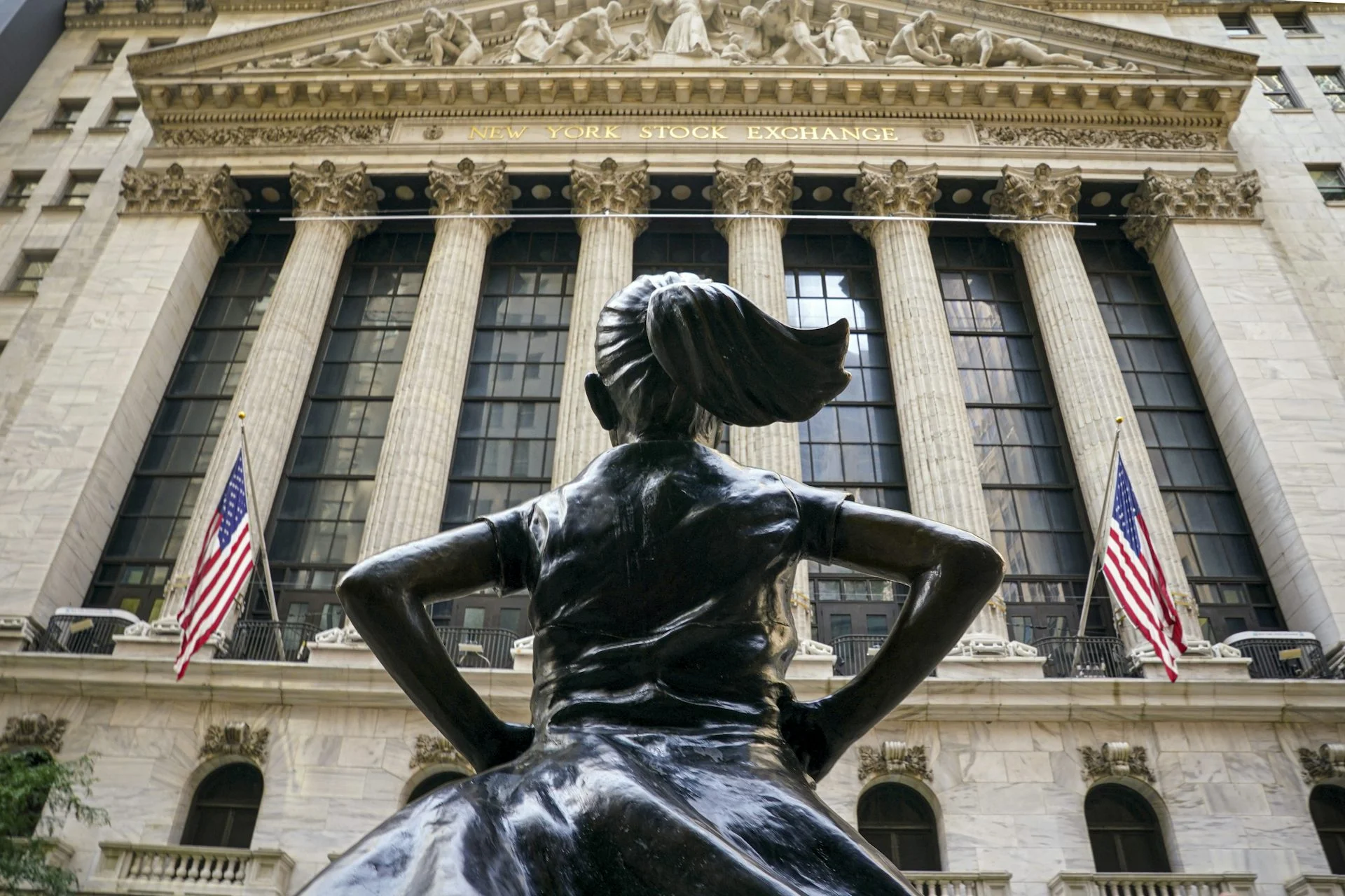A statue of a girl with a ponytail, hands on her hips and seen from behind, is positioned in front of a building with large pillars.