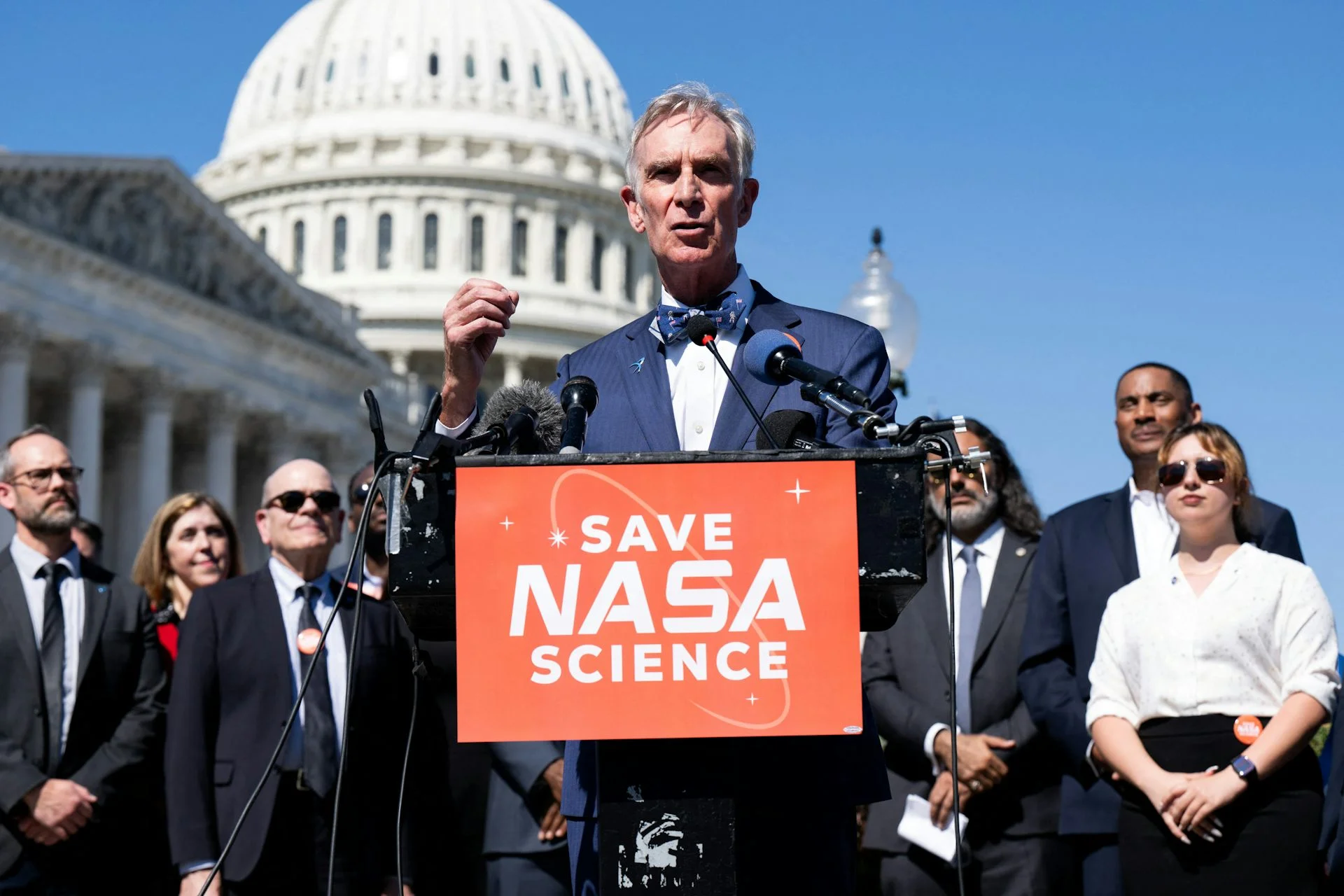 A white older man wearing a blue blazer and a bow tie stands at a podium that says 'Save NASA Science' in front of the US Capitol building, with a group of people dressed formally standing behind him.