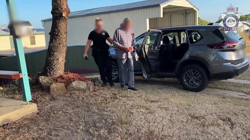 A man with handcuffs on being walked to a car with an open door, near a home.