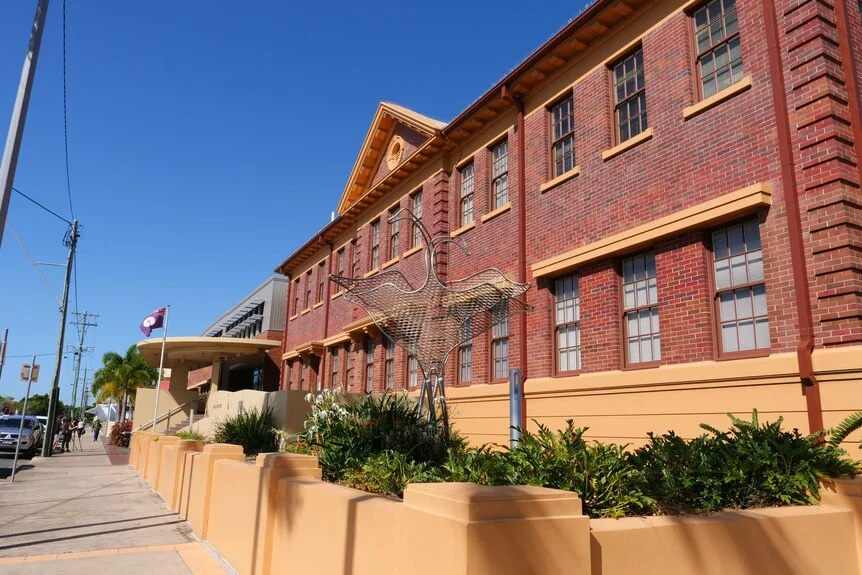 Anj old brick building on a sunny day with a bird statue out the front and a Queensland flag flying.