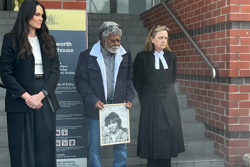 An Indigenous man with grey hair and beard holds a picture as he stands beside a judge in robes.