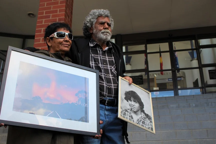 Two Aboriginal elders stand on the front of courthouse steps holding each other and the photo of their dead family member.