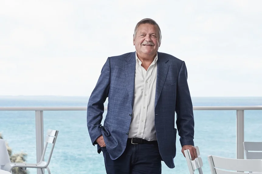 A middle aged white man with short grey hair standing on decking by the ocean