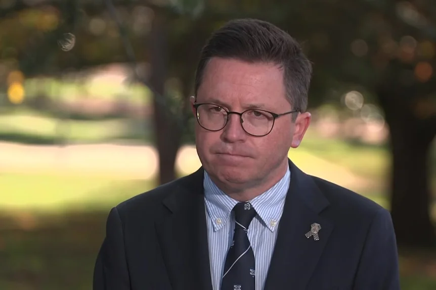 A man with dark hair and glasses in a navy jacket, striped shirt and dark tie stands in a park.