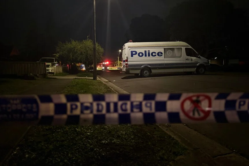 A dark night scene with police tape at the front and a police car in the back.