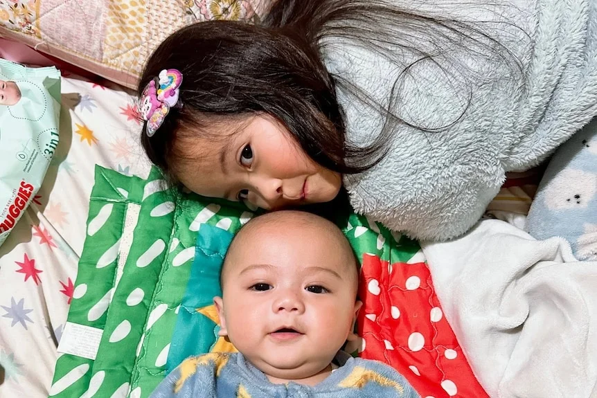 A young Asian girl with a grey sweater, smiling on a bed next to her baby brother smiling.