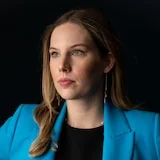 A woman with dark blonde hair and a blue jacket looks to the side with serious expression.