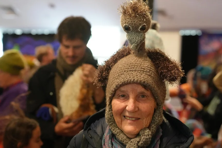 An older woman looking at the camera, wearing a brown beanie with a big emu character knitted to its top.