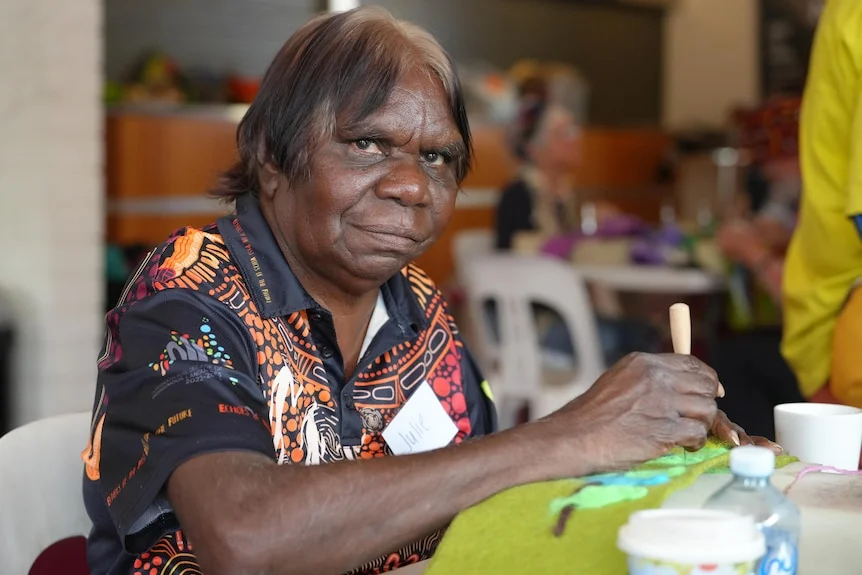A mid-shot of an Aboriginal woman, with show gray / brown hair, sitting with a felt needle in hand, looking at camera.