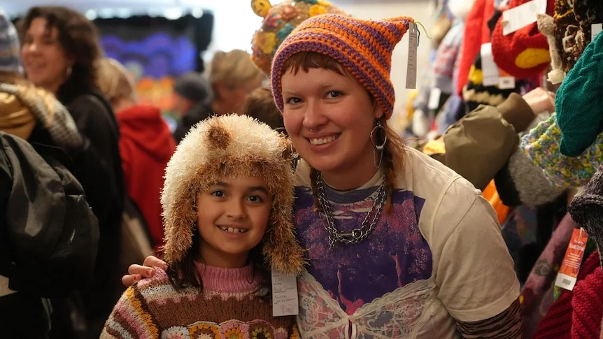 A young girl wearing a fluffy brown beanie and young woman with a striped purple / orange beanie standing close in crowd.