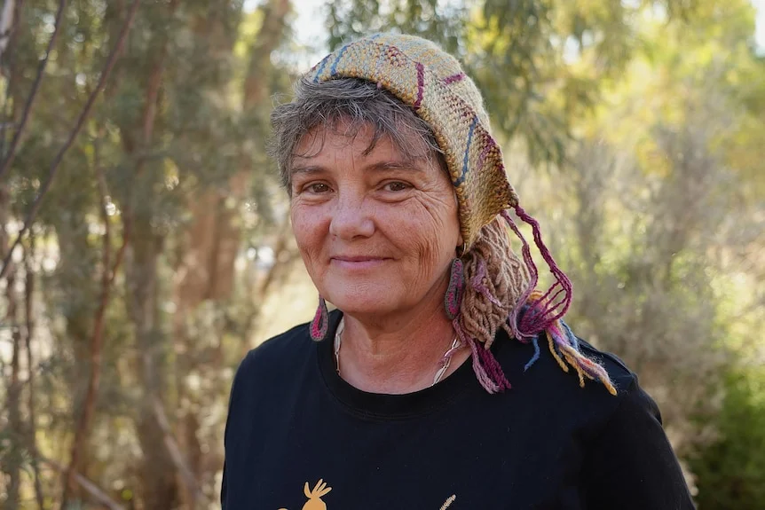 Jo smiling at the camera, she is wearing a colourful knitted beanie, forest behind her.
