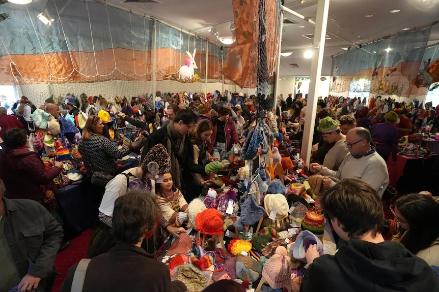 A big crowd of people in an indoor large room, most wearing beanies, looking over beanie products laid out on table stalls.