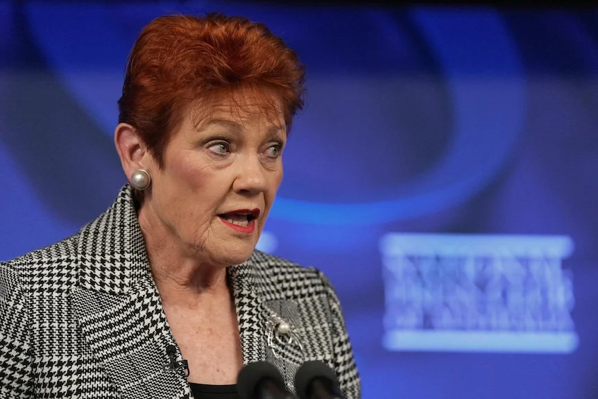 A woman with red hair, wearing a checked coat, standing in front of a blue background.