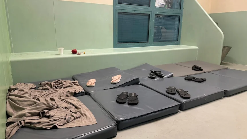 A watch house cell with multiple pairs of thongs lined up on mattresses on the floor. 