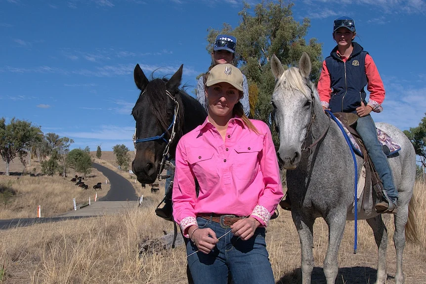 A woman in a pink shirt stands in front of two girls on horses.
