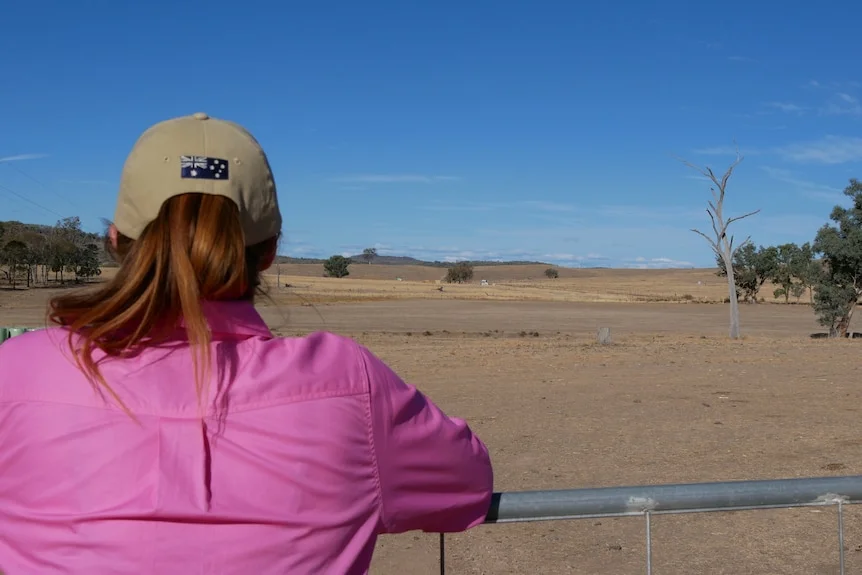 A woman in a pink shirt and cap looking out over a brown paddock.