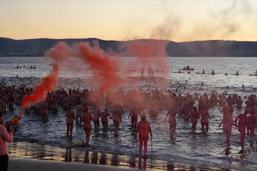 a crowd of naked people run into the river as red flares are shot into the air.
