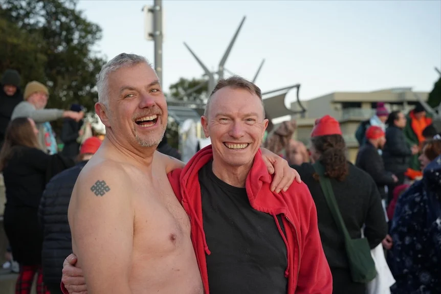 two men stand with arms wrapped around each other, smiling.
