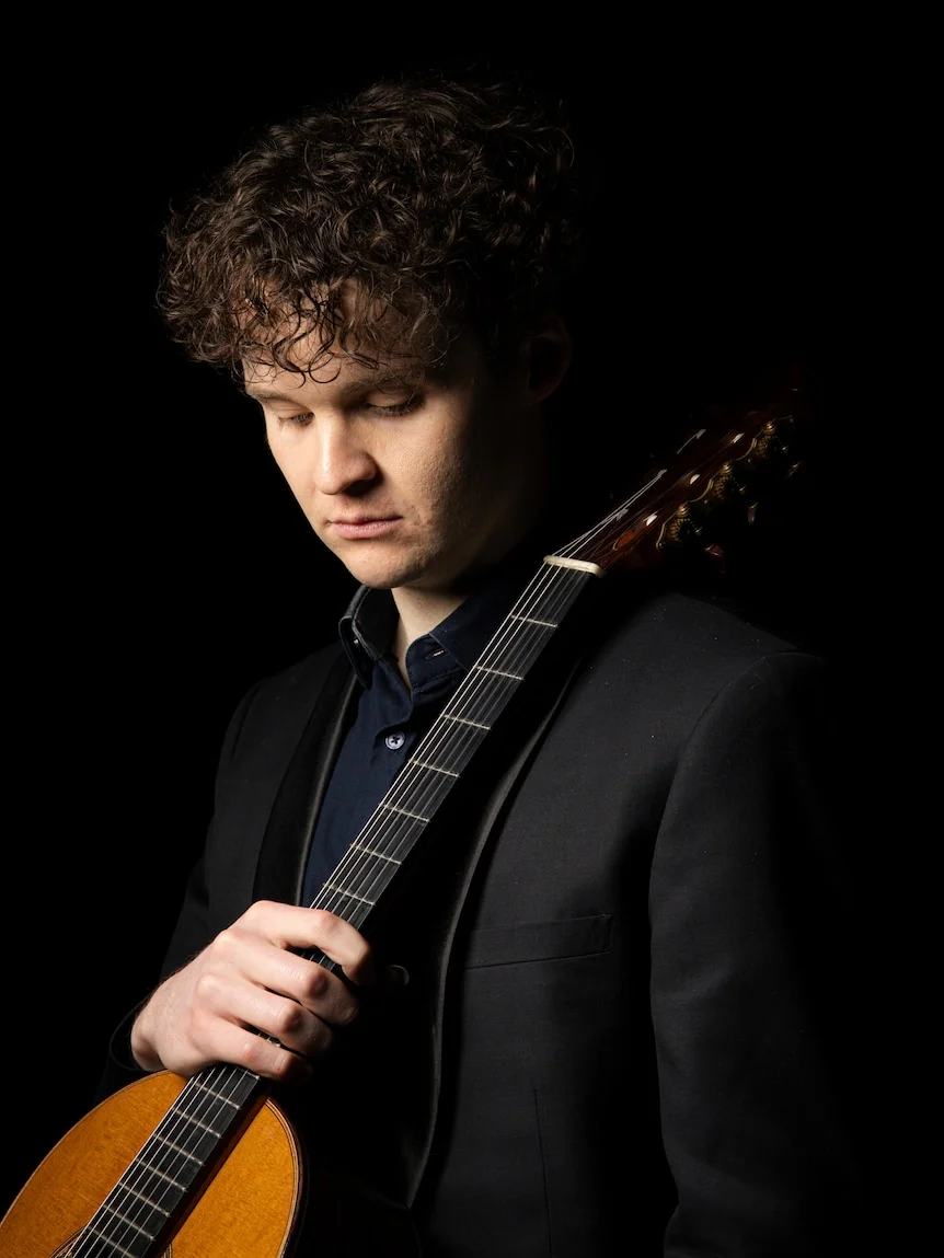 Studio portrait of curly-haired classical guitarist, Connor Whyte holding instrument against shoulder, on a black background.