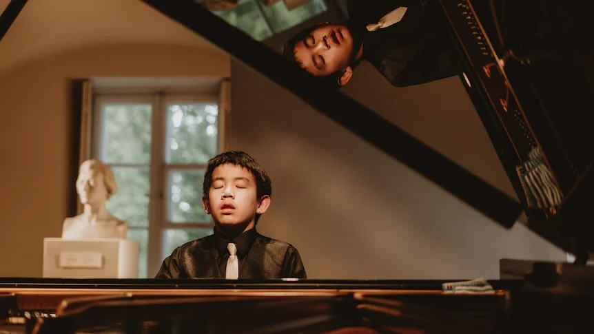 Dillon Chan seated at a grand piano with eyes closed, his reflection visible on lid, with a composer bust behind.