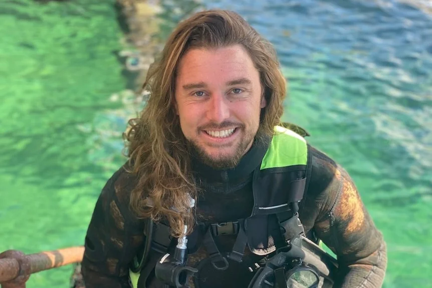 A man in a surfer wetsuit carrying a large camera, light equipment and flippers while walking up step from the ocean