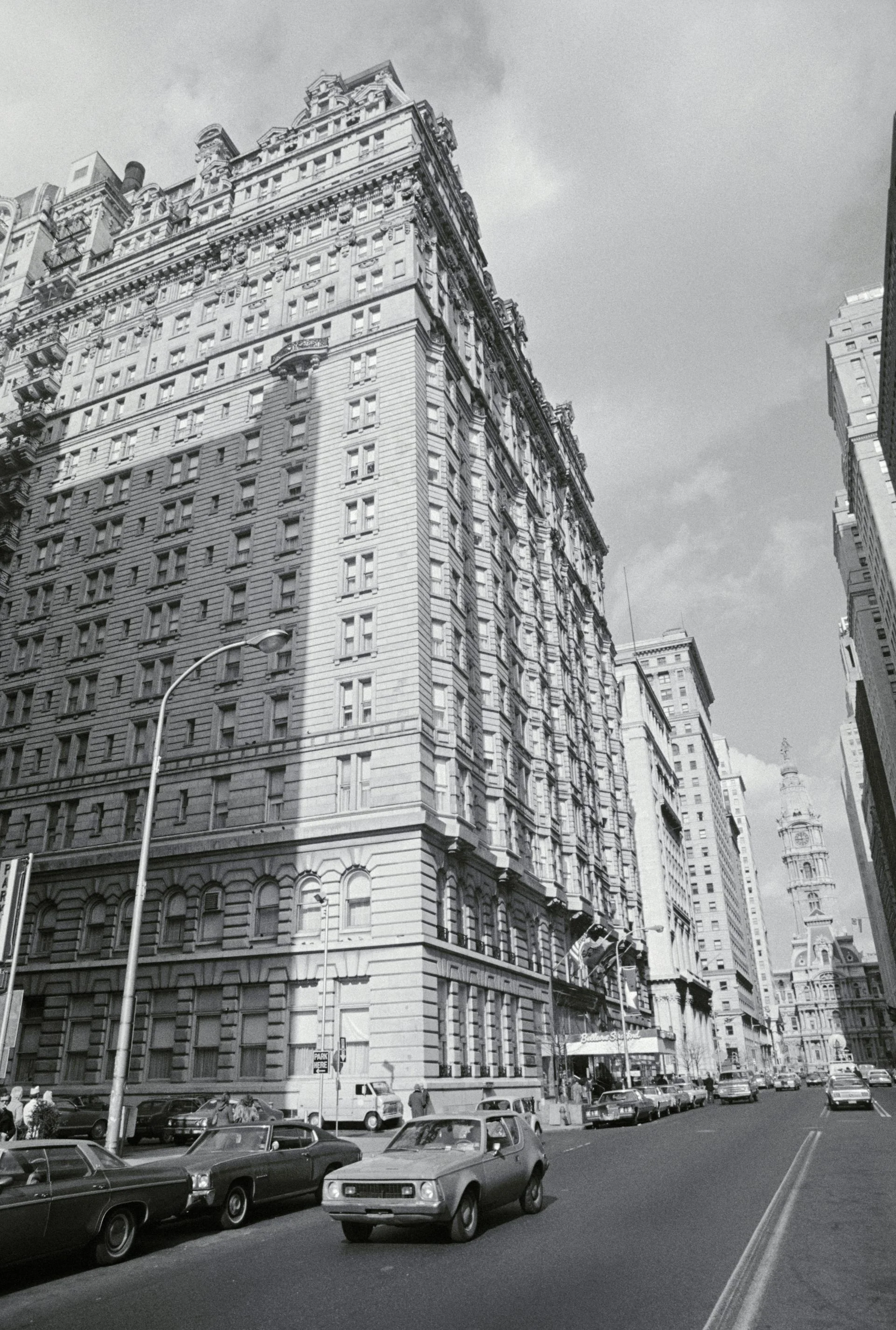 Black-and-white photo of large ornate building alongside two-way street with 1970s-era cars