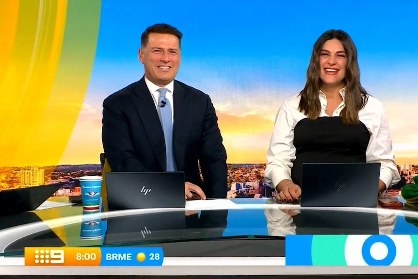 Karl wears a suit and sits at a desk in a television studio. A smiling woman sits next to him.