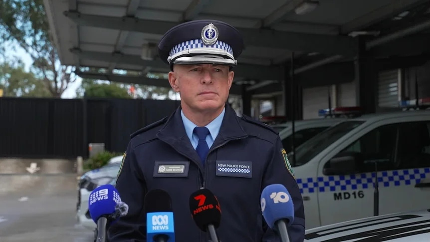 Darrin wears a police hat and uniform and speaks into media microphones.