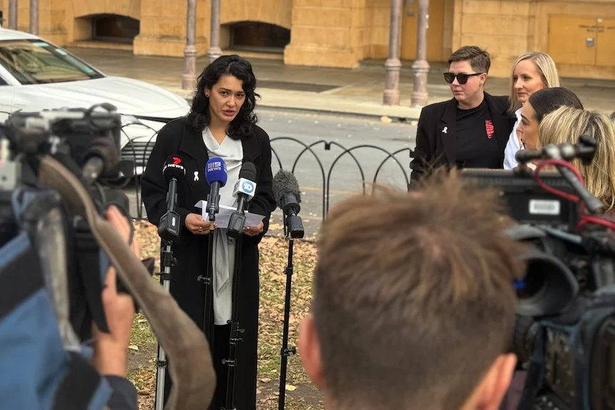 A woman holds a paper and speaks into media microphones while being filmed and watched on by reporters