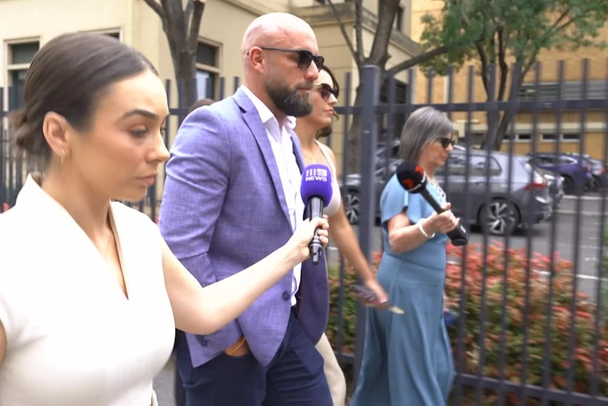 A man in a purple blazer leaves a court building surrounded by a media scrum