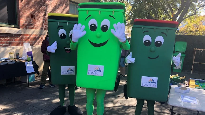 A green fogo bin mascot next to a recycling bin mascot and a landfill bin mascot outside near trees.