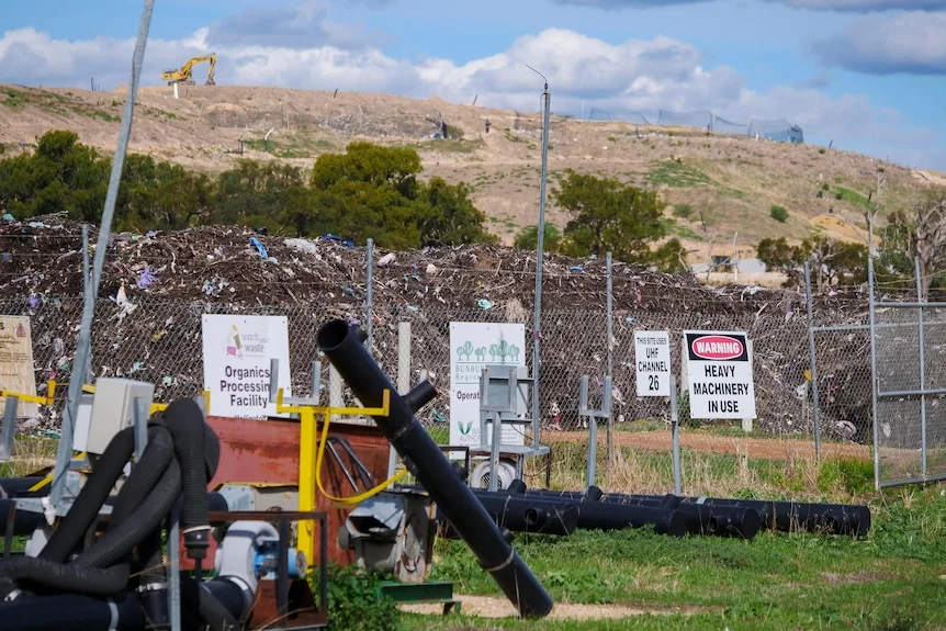 Large piles of soil with bits of rubbish throughout behind a metal fence. In the distance, an excavator sits on a hill.