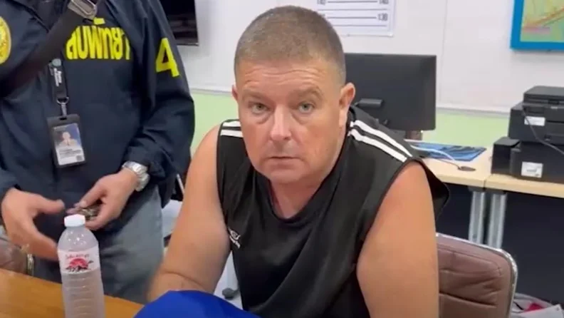 A man sits in a singlet behind a desk looking at the camera