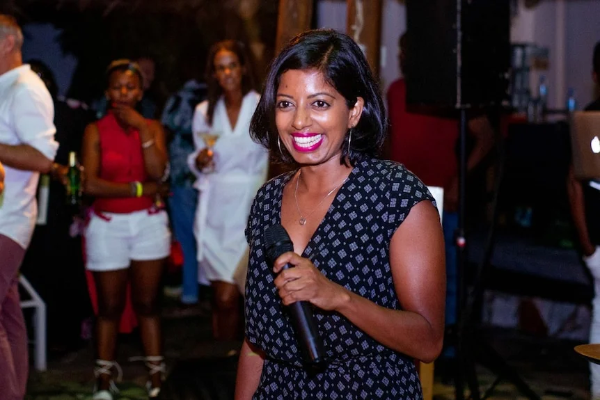 Sashi Perera stands with a microphone in her hand, smiling. It is night time and there are people in the background.
